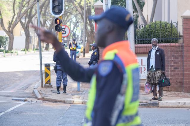 A pedestrian (R) waits to cross a street as South African Police Service (SAPS) officers (L) and a Johannesburg Metro Police Department (JMPD) officer (C) secures a road leading to the Nasrec Expo Centre, in Rosebank, Johannesburg on November 22, 2025, ahead of the opening of the G20 Leaders’ Summit. (Photo by Zinyange Auntony / AFP)