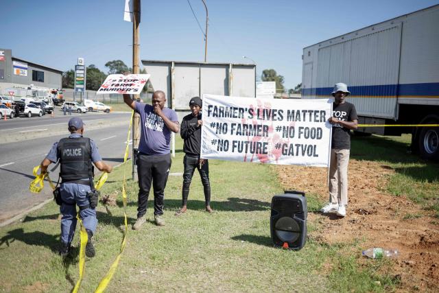 A South African Police Service (SAPS) officer (L) walks towards Farmer Lives Matter protesters holding placards next to a road near the Nasrec Expo Centre in Johannesburg on November 22, 2025 ahead of the opening of the G20 Leader's Summit. (Photo by EMMANUEL CROSET / AFP)