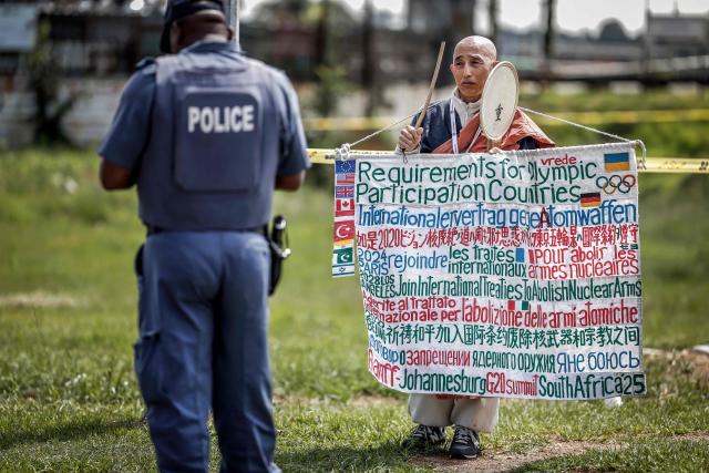 A South African Police Service (SAPS) officer (L) stands near Japanese Buddhist Monk and Anti-nuclear campaigner Toyoshige Sekiguchi (R) who is protesting next to a road near the Nasrec Expo Centre in Johannesburg on November 22, 2025 ahead of the opening of the G20 Leaders' Summit. (Photo by Marco Longari / AFP)