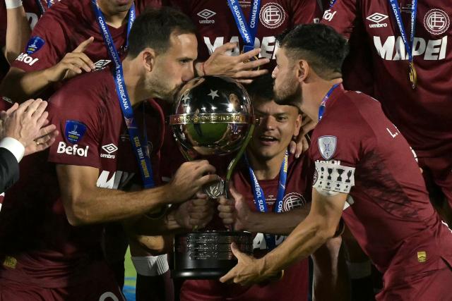 TOPSHOT - Lanus' defender #24 Carlos Izquierdoz (L) kisses the trophy next to forwards #07 Lautaro Acosta (R) and #09 Walter Bou (C) after winning the Copa Sudamericana final football match between Argentina's Lanus and Brazil's Atletico Mineiro at the Defensores del Chaco stadium in Asuncion on November 22, 2025. (Photo by Juan MABROMATA / AFP)