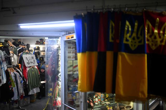 A vendor sits in a souvenir kiosk next to a doormat bearing an image depicting Russia's President displayed among other items in Kyiv on November 23, 2025, amid the Russian invasion of Ukraine. (Photo by Sergei GAPON / AFP)