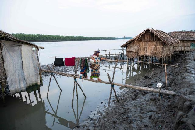 In this photograph taken on July 12, 2025, Lipika Mondal, a climate-displaced woman walks past stilt houses with a water pot as she crosses a bamboo bridge over Shibsa river at New Jhulonto Para, a village vulnerable to rising sea levels in Khulna district. On Bangladesh's coast, where mighty Himalayan rivers meet the sea, water defines every rhythm of life and every struggle. For the millions living in the ecologically sensitive deltas of mudflats and mangrove forests, finding clean drinking water has become an escalating challenge. Rising seas driven by climate change are swallowing low-lying areas, while stronger storms push saltwater further inland, turning wells and lakes brackish, according to government scientists. (Photo by Muhammad Amdad HOSSAIN / AFP) / To go with 'BANGLADESH-ENVIRONMENT-CLIMATE-WATER, REPORTAGE' by Muhammad Amdad HOSSAIN