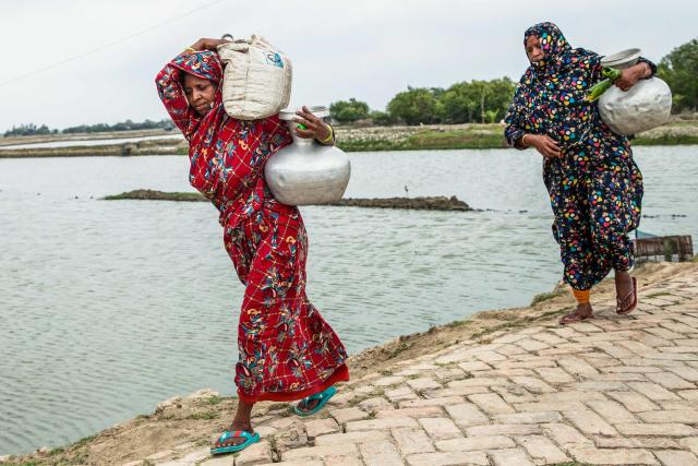 In this photograph taken on April 25, 2025, women walk home after fetching pots of water from the Kholpetua river at Parshemari village in Khulna district. On Bangladesh's coast, where mighty Himalayan rivers meet the sea, water defines every rhythm of life and every struggle. For the millions living in the ecologically sensitive deltas of mudflats and mangrove forests, finding clean drinking water has become an escalating challenge. Rising seas driven by climate change are swallowing low-lying areas, while stronger storms push saltwater further inland, turning wells and lakes brackish, according to government scientists. (Photo by Muhammad Amdad HOSSAIN / AFP) / To go with 'BANGLADESH-ENVIRONMENT-CLIMATE-WATER, REPORTAGE' by Muhammad Amdad HOSSAIN
