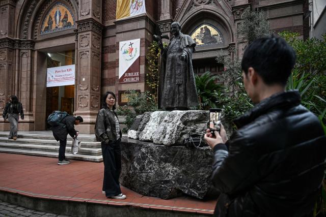 A woman poses for a photo in front of the statue of late pope Jean XXIII outside the Saint Anthony of Padua Church as a welcome banner bearing a portrait of Pope Leo XIV is displayed on its gate, ahead of his visit to Turkey, in Istanbul on November 24, 2025. (Photo by Ozan KOSE / AFP)