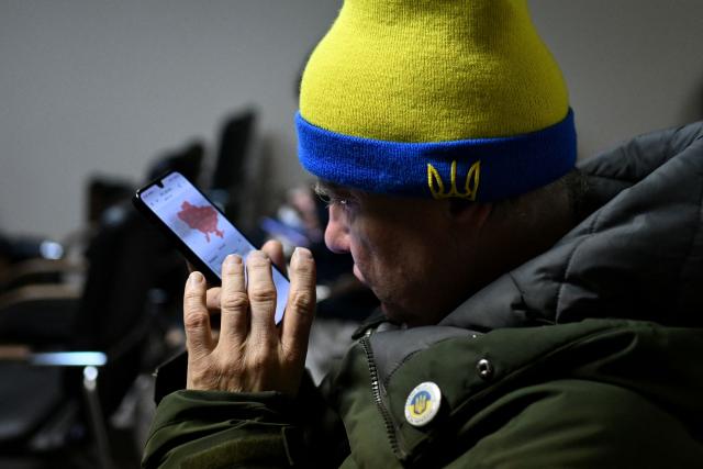 A man wearing a hat in the colours of the Ukrainian flag with the embroidered Ukrainian coat of arms, checks the air raid alert map on his mobile phone as he sits in the hotel's underground shelter during an air attack in Kyiv early on November 25, 2025, amid the Russian invasion of Ukraine. Powerful explosions rocked Ukraine's capital Kyiv, an AFP journalist heard, as the air force issued a countrywide missile warning early November 25, 2025. (Photo by Sergei GAPON / AFP)