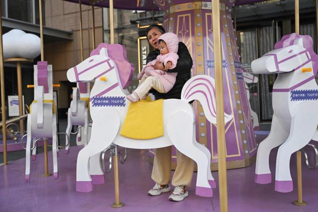A woman accompanies a toddler on a carousel ride in front of a shopping mall in Beijing on November 25, 2025. (Photo by Pedro PARDO / AFP)
