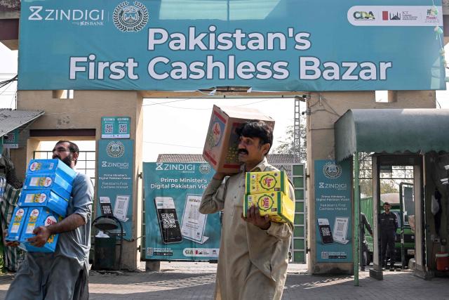 Shopkeepers carry goods past digital cashless payment QR (quick response) codes displayed at a market in Islamabad on November 25, 2025. (Photo by Farooq NAEEM / AFP)