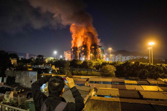 An onlooker takes photos as a major fire engulfs several apartment blocks at the Wang Fuk Court residential estate (background) in Hong Kong's Tai Po district on November 26, 2025. At least four people were killed when a fire engulfed several high-rise blocks in a Hong Kong residential estate on November 26, the government said, with media reporting that some residents were trapped inside. (Photo by AFP)