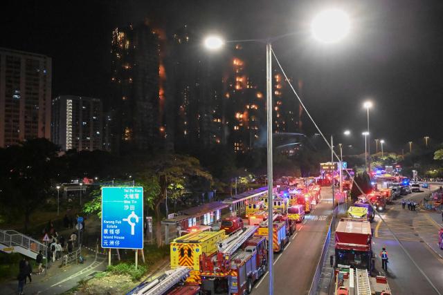 Fire trucks and rescue vehicles line a road during a major fire at the Wang Fuk Court residential estate (background) in Hong Kong's Tai Po district on November 26, 2025. At least four people were killed when a fire engulfed several high-rise blocks in a Hong Kong residential estate on November 26, the government said, with media reporting that some residents were trapped inside. (Photo by Peter PARKS / AFP)
