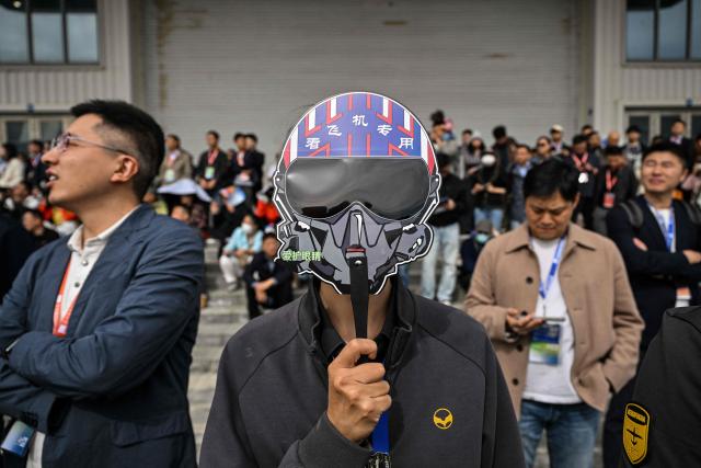 A woman holds a mask resembling a pilot's helmet as she watches an aerobatic performance during the Aero Asia 2025 in Zhuhai, in southern China's Guangdong province on November 27, 2025. Aero Asia 2025 is an international aviation and aerospace exhibition that runs between November 27 and 30. (Photo by Hector RETAMAL / AFP)