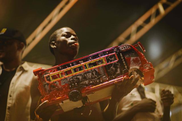 A member of Kenyan artist Parroty Vunulu’s team holds a miniature matatu -Kenya’s iconic, privately owned public transport minibus- during a performance for the 4th Nganya Awards  in Nairobi on November 29, 2025. The Nganya Awards is a celebration of Kenya’s matatu culture, honoring the matatu scene and the unique lifestyle around them. This years awards had over 40 categories, from matatu crews to vehicle designers, sound system and social influencers linked to the matatu lifestyle, with voting taking place ahead of the award show. (Photo by Fredrik LERNERYD / AFP)