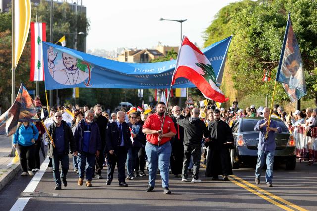 Faithful march carrying the Lebanese flag and a banner as others wait for Pope Leo XIV in Beirut's eastern suburb of Hazmieh, where the Pope will drive past as he makes his way to the Presidential Palace on November 30, 2025. Pope Leo XIV wrapped up a four-day trip to Turkey on November 30, 2025, after a warm welcome by its tiny Christian community, before heading to Lebanon with a message of peace for the crisis-mired nation. (Photo by AFP)
