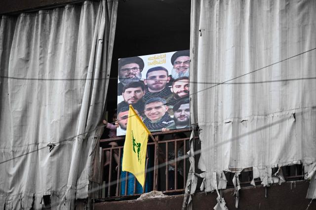 People stand on a balcony decorated with a Hezbollah flag and holding a placard with the images of Hezbollah's assassinated leaders and others as they wait for the arrival of Pope Leo XIV in Beirut's southern suburbs, a packed residential area known as Dahiyeh, which is also a Hezbollah bastion, on November 30, 2025. Pope Leo XIV arrived in Lebanon for a two-day trip where he is expected to bring a message of peace, an AFP journalist aboard the plane said. (Photo by JOSEPH EID / AFP)