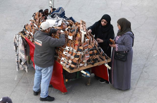 Vendors of traditional Marrakech shoes speak to a customer in Jemaa el-Fna Square or Jamaa Lafna, listed as UNESCO Cultural Heritage site, located in the heart of the Medina of the city of Marrakech on November 30, 2025. (Photo by Abdel Majid BZIOUAT / AFP)