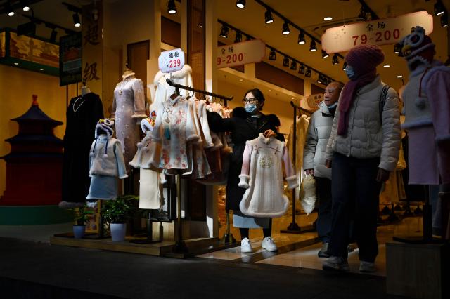 An employee (L) pitches clothes to customers walking past at a clothing shop in Beijing on December 2, 2025. (Photo by WANG Zhao / AFP)