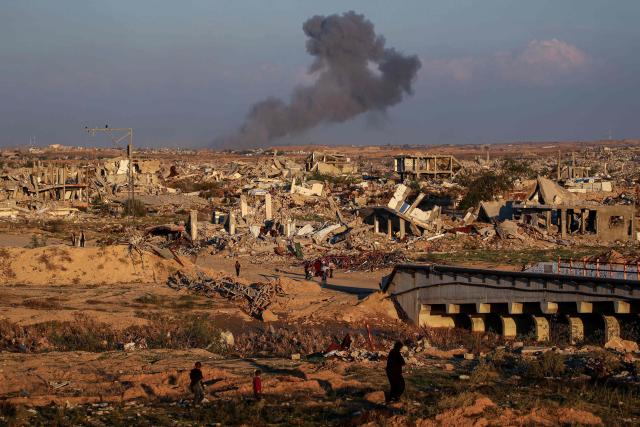 TOPSHOT - People walk amid the rubble of destroyed buildings at the Nuseirat camp for displaced Palestinians as smoke billows in the distance following Israeli strikes east of Gaza City on December 2, 2025. A ceasefire which came into effect on October 10 remains fragile, with Israel and Hamas accusing each other of violating the terms, while the Gaza Strip remains in a deep humanitarian crisis. (Photo by Eyad Baba / AFP)