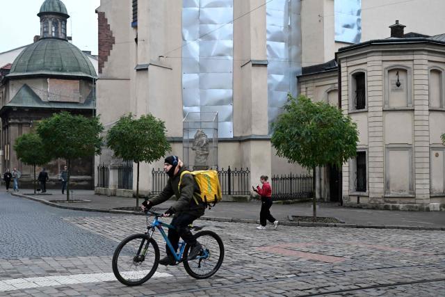 A cyclist working for the food delivery service Glovo rides a bicycle past the Latin Cathedral, with its windows protected by metal plates and statue encased in a protective structure against possible damage caused by air strikes, in Lviv on December 2, 2025, amid the Russian invasion of Ukraine. (Photo by Sergei GAPON / AFP)