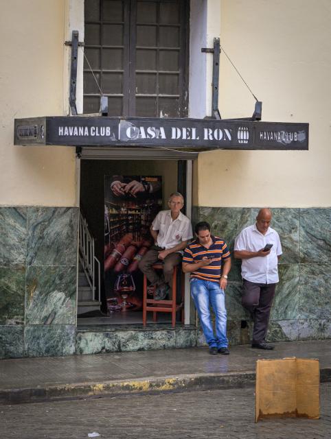 Three workers at a state-run resort sit and check their phones on the sidewalk during a power outage in Havana, on December 3, 2025. Millions of people were left without electricity on December 3 in western Cuba, including Havana, due to damage to the power grid in that region of the island, which has been suffering recurrent blackouts for two years. (Photo by ADALBERTO ROQUE / AFP)