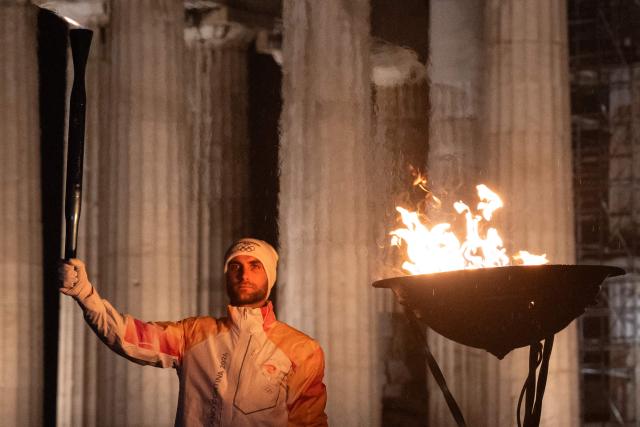 Greek rower Stefanos Ntouskos holds the Olympic flame in front of the ancient temple of the Parthenon atop the Acropolis hill during the Olympic torch relay in Athens on December 3, 2025. (Photo by Angelos TZORTZINIS / AFP)