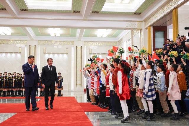 China's President Xi Jinping (L) and France's President Emmanuel Macron greet children waving Chinese flags and holding up flowers during a welcome ceremony at the Great Hall of the People in Beijing on December 4, 2025. (Photo by Sarah Meyssonnier / POOL / AFP)