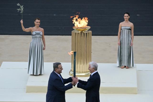 Hellenic Olympic Committee President Isidoros Kouvelos (L) and President of the Italian National Olympic Committee and CEO of the Milan Cortina 2026 Winter Olympic Giovanni Malago (R) hand the torch next to Greek actress Mary  Mina playing the role of the High Priestess, during the handover Olympic flame ceremony for the Milano-Cortina 2026 Winter Olympic Games and Paralympic Games, at the Panathinean stadium in Athens, on December 4, 2025. (Photo by Angelos Tzortzinis / AFP)