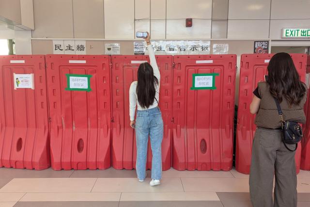 A woman takes a photo with her phone above barricades in front of the student union-run notice board, nicknamed “democracy wall”, at Hong Kong Baptist University in Hong Kong on December 3, 2025. A Hong Kong university ordered its student union to shut down after a message was posted on campus expressing condolences and urging justice for the victims of a major fire, according to a letter publicly shared on December 5. A message expressing condolences and urging justice for the victims in a deadly fire at Wang Fuk Court was seen on the notice board before it was blocked off. (Photo by Holmes CHAN / AFP)