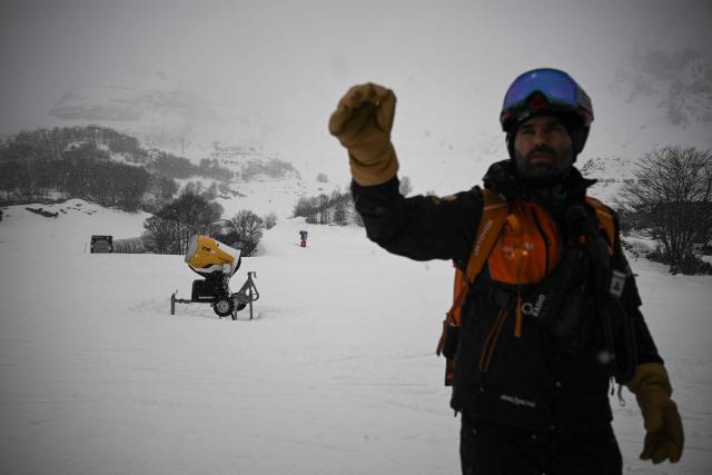 A ski patroller stands next a tower-based snow gun system used at ski resorts to produce artificial snow for ski slopes and winter sports facilities, at the Gourette ski resort near the Col d'Aubisque pass in the Pyrenees, south-western France on December 2, 2025. The family-oriented resort features one of the largest beginner areas in the Pyrenees and is a pioneer in the mountain range where skiing was first introduced in 1903 near Col d'Aubisque pass. (Photo by Philippe LOPEZ / AFP)