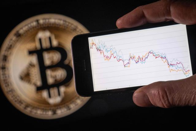 This illustration photograph shows a man holding a phone with a downward trend in the stock market in front of a screen displaying a Bitcoin logo, in Mulhouse on December 5, 2025. (Photo by SEBASTIEN BOZON / AFP)