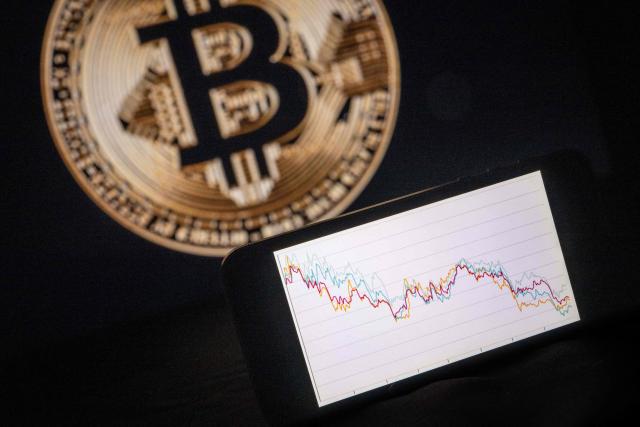 This illustration photograph shows a phone with a downward trend in the stock market in front of a screen displaying a Bitcoin logo, in Mulhouse on December 5, 2025. (Photo by SEBASTIEN BOZON / AFP)