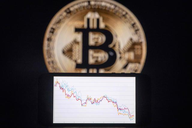 This illustration photograph shows a phone with a downward trend in the stock market in front of a screen displaying a Bitcoin logo, in Mulhouse on December 5, 2025. (Photo by SEBASTIEN BOZON / AFP)