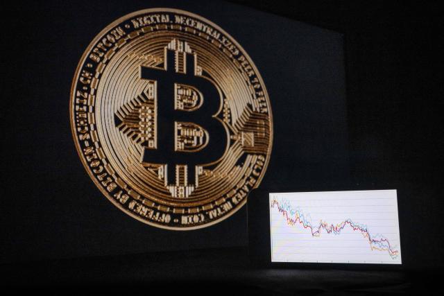 This illustration photograph shows a phone with a downward trend in the stock market in front of a screen displaying a Bitcoin logo, in Mulhouse on December 5, 2025. (Photo by SEBASTIEN BOZON / AFP)