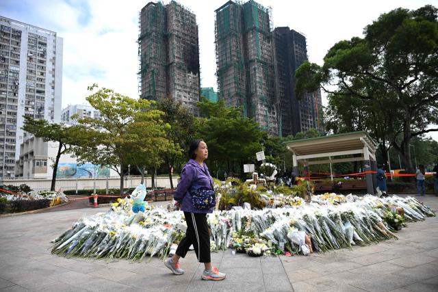 A woman walks past flowers laid for the victims of a deadly fire at the Wang Fuk Court residential estate in Tai Po district of Hong Kong on December 7, 2025. Hong Kong began voting on December 7 to choose new lawmakers under Beijing's "patriots only" rules, though government efforts to drive turnout have been overshadowed by the city's deadliest fire in decades. (Photo by Peter PARKS / AFP)