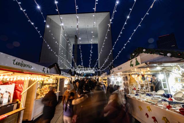 TOPSHOT - Visitors walk and shop at the Christmas market on the Parvis de La Defense, with La Grande Arche of La Defense in the background, at the business district of La Defense, in Puteaux outside Paris, on December 7, 2025. (Photo by Ian LANGSDON / AFP)