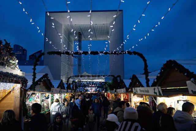 Visitors walk and shop at the Christmas market on the Parvis de La Defense, with La Grande Arche of La Defense in the background, at the business district of La Defense, in Puteaux outside Paris, on December 7, 2025. (Photo by Ian LANGSDON / AFP)