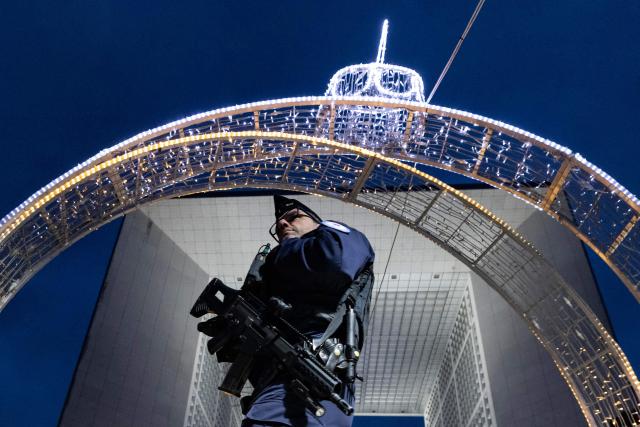 A French Republican Security Corps (CRS - Compagnies Republicaines de Securite) police officer patrols inside the Christmas market on the Parvis de La Defense, with the Grande Arche de la Defense in the background at the business district of La Defense, in Puteaux outside Paris, on December 7, 2025. (Photo by Ian LANGSDON / AFP)