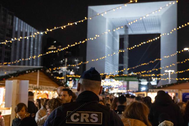 A French Republican Security Corps (CRS - Compagnies Republicaines de Securite) police officer patrols inside the Christmas market on the Parvis de La Defense, with the Grande Arche de la Defense in the background at the business district of La Defense, in Puteaux outside Paris, on December 7, 2025. (Photo by Ian LANGSDON / AFP)