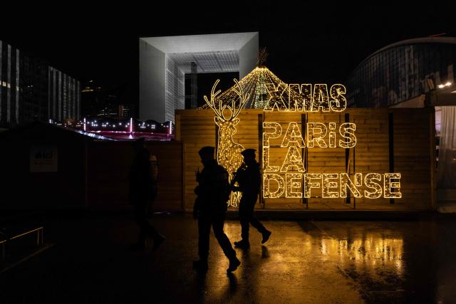 French Republican Security Corps (CRS - Compagnies Republicaines de Securite) police officers patrol past illuminated signage around the Christmas market on the Parvis de La Defense, with La Grande Arche of La Defense in the background, at the business district of La Defense, in Puteaux outside Paris, on December 7, 2025. (Photo by Ian LANGSDON / AFP)