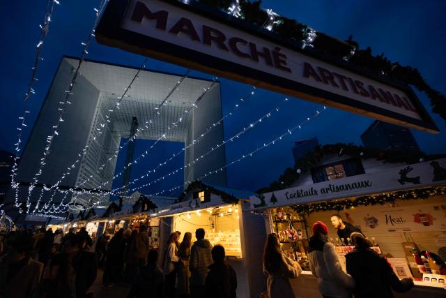 Visitors walk and shop at the Christmas market on the Parvis de La Defense, with La Grande Arche of La Defense in the background, at the business district of La Defense, in Puteaux outside Paris, on December 7, 2025. (Photo by Ian LANGSDON / AFP)