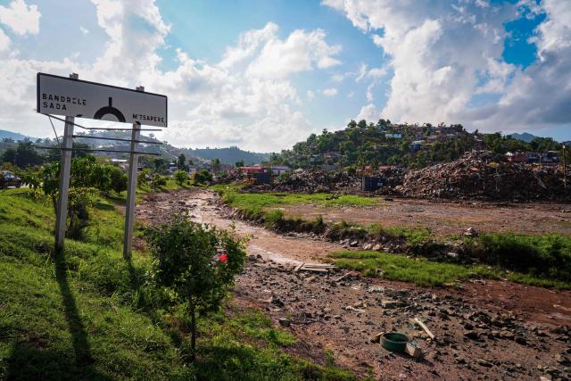 This photograph shows a plot of land filled with debris from houses and metal sheets  with a road sign indicating the village of M'tsapere following the 2024 Cyclone Chido, in Mamoudzou, on the French Indian Ocean territory of Mayotte, on December 2, 2025. Chido, the most devastating cyclone to hit Mayotte in the last 90 years, hit France's poorest department on December 14, 2024, causing colossal damage. (Photo by Marine GACHET / AFP)
