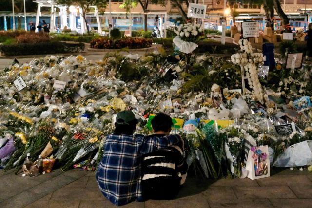 Volunteers sit in front of the flowers left by mourners for the victims of a deadly fire at the Wang Fuk Court residential estate in Tai Po district of Hong Kong on December 8, 2025. Hong Kong's leader said on December 8 that the fresh crop of lawmakers who will take office after the "patriots only" legislative election will drive institutional reform, following the city's deadliest fire in decades. (Photo by Tommy WANG / AFP)
