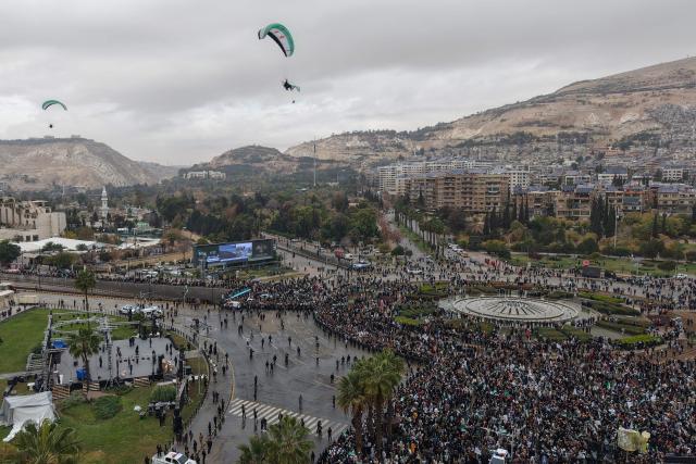 An areal photograph shows paragliders flying over people celebrating a year since the ousting of longtime ruler Bashar al-Assad in the Syrian capital Damascus on December 8, 2025. President Ahmed al-Sharaa on December 8 urged Syrians to work together to rebuild their country as they marked a year since the ousting of longtime ruler Bashar al-Assad. Sharaa's Islamist-led alliance launched a lightning offensive in late November last year, taking the capital Damascus on December 8 after nearly 14 years of war and putting an end to more than five decades of the Assad family's iron-fisted rule. (Photo by Bakr ALKASEM / AFP)