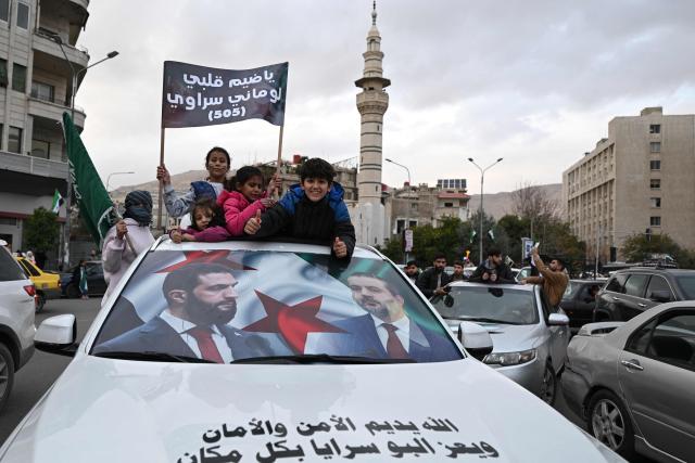 People ride a car reflecting the faces of Syria's President Ahmed al-Sharaa and Syrian Minister of Foreign Affairs and Expatriates Asaad Al-Shaibani as they celebrate a year since the ousting of longtime ruler Bashar al-Assad in the Syrian capital Damascus on December 8, 2025. President Ahmed al-Sharaa on December 8 urged Syrians to work together to rebuild their country as they marked a year since the ousting of longtime ruler Bashar al-Assad. Sharaa's Islamist-led alliance launched a lightning offensive in late November last year, taking the capital Damascus on December 8 after nearly 14 years of war and putting an end to more than five decades of the Assad family's iron-fisted rule. (Photo by LOUAI BESHARA / AFP)