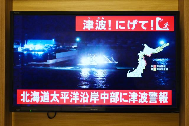 TOPSHOT - An advisory reading "Tsunami! Run!" (top) and "Tsunami warning issued for central Hokkaido’s Pacific Coast" flashes over the live footage of a boat leaving a port, on a television screen in Sapporo, in Japan’s northern Hokkaido prefecture on December 8, 2025. Japan recorded a 40-centimetre (16-inch) tsunami after a 7.6-magnitude quake on December 8. (Photo by GREG BAKER / AFP)