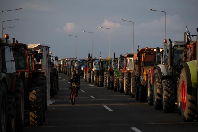 A farmer rides a bicycle between parked tracktors blocking a national highway outside the central Greek city of Karditsa on December 8, 2025. Greek farmers demanding the payment of EU subsidies on December 8 shut down the two main international airports on the island of Crete after clashing with riot police in a growing showdown with the government. (Photo by Aris MESSINIS / AFP)