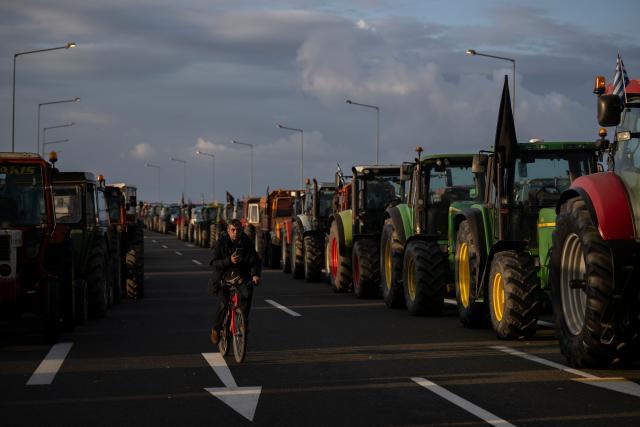 A farmer rides a bicycle between parked tracktors blocking a national highway outside the central Greek city of Karditsa on December 8, 2025. Greek farmers demanding the payment of EU subsidies on December 8 shut down the two main international airports on the island of Crete after clashing with riot police in a growing showdown with the government. (Photo by Aris MESSINIS / AFP)