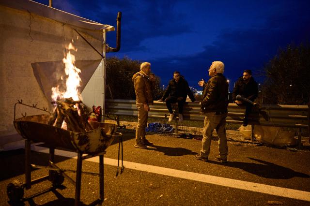 Farmers chat next to a fire as they block the national highway with their tractors, outside the central Greek city of Karditsa on December 8, 2025 to demand swifter access to EU subsidies. Greek farmers demanding the payment of EU subsidies on December 8 shut down the two main international airports on the island of Crete after clashing with riot police in a growing showdown with the government. (Photo by Aggelos NAKKAS / AFP)