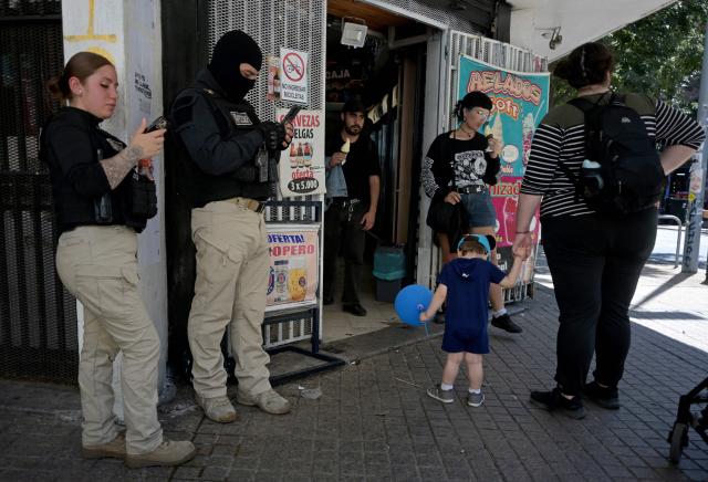 Municipal security agents check their phones as people line up at an ice-cream shop at the Lastarria neighborhood in Santiago on December 8, 2025. Chile will choose its next president in a runoff on December 14, with far-right candidate Jose Antonio Kast as the clear favorite against leftist Jeannette Jara, after a campaign marked by fears over crime and irregular migrants. (Photo by Eitan ABRAMOVICH / AFP)