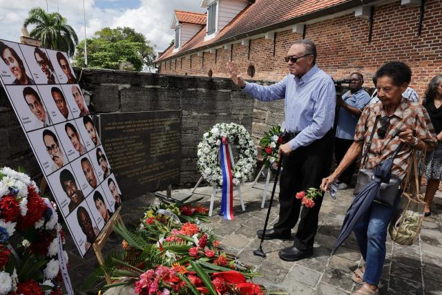 A relative of the 15 victims of the December 1982 murders at the former Fort Zeelandia military barracks lies flowers in front of their pictures during a remembrance ceremony in Paramaribo on December 8, 2025. Relatives of the 15 victims killed filed a lawsuit against the government demanding rehabilitation and compensation. (Photo by Ranu ABHELAKH / AFP)
