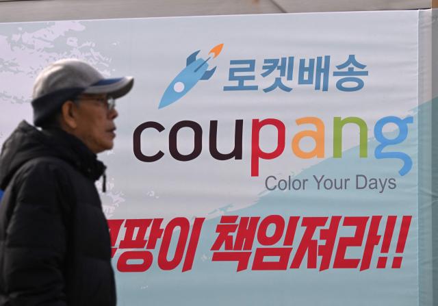 A man walks past a banner showing the logo of South Korean online delivery service Coupang, displayed by transportation union members to demand workers' rights outside a building housing the headquarters of Coupang in Seoul on December 9, 2025. South Korean police raided the Seoul headquarters of e-commerce giant Coupang on December 9, over a recent data leak believed to have affected almost two-thirds of the country's population. (Photo by Jung Yeon-je / AFP)
