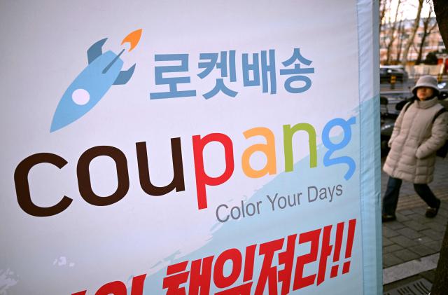 A woman walks past a banner showing the logo of South Korean online delivery service Coupang, displayed by a transport workers' union to demand better labour rights outside a building housing the headquarters of Coupang in Seoul on December 9, 2025. South Korean police raided the Seoul headquarters of e-commerce giant Coupang on December 9, over a recent data leak believed to have affected almost two-thirds of the country's population. (Photo by Jung Yeon-je / AFP)